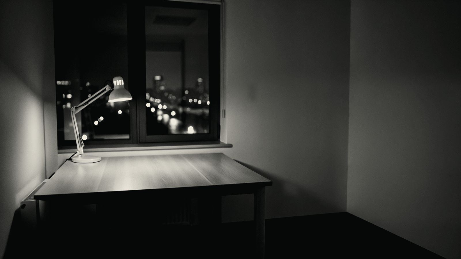 An empty office desk at night lit by a single lamp with a laptop screen glowing, suggesting work still running after everyone has gone home