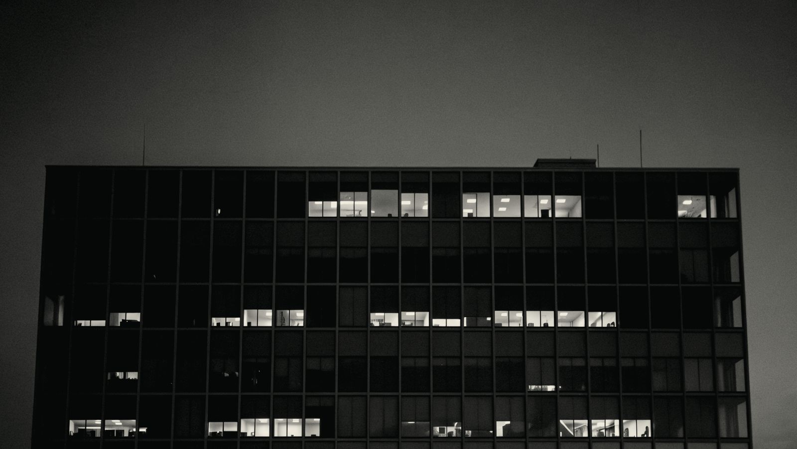 An office lit warmly at night seen through its windows, an image of work still going on inside systems long after the people have gone home
